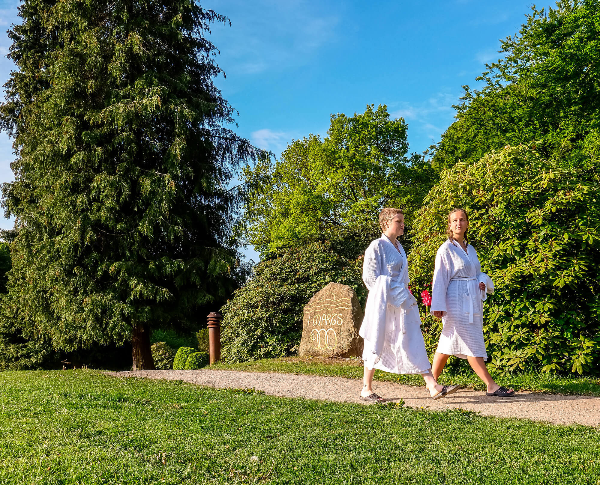 Kvinder i badekåber går på en sti i en park med en mindesten for Vejlefjords grundlæggelse