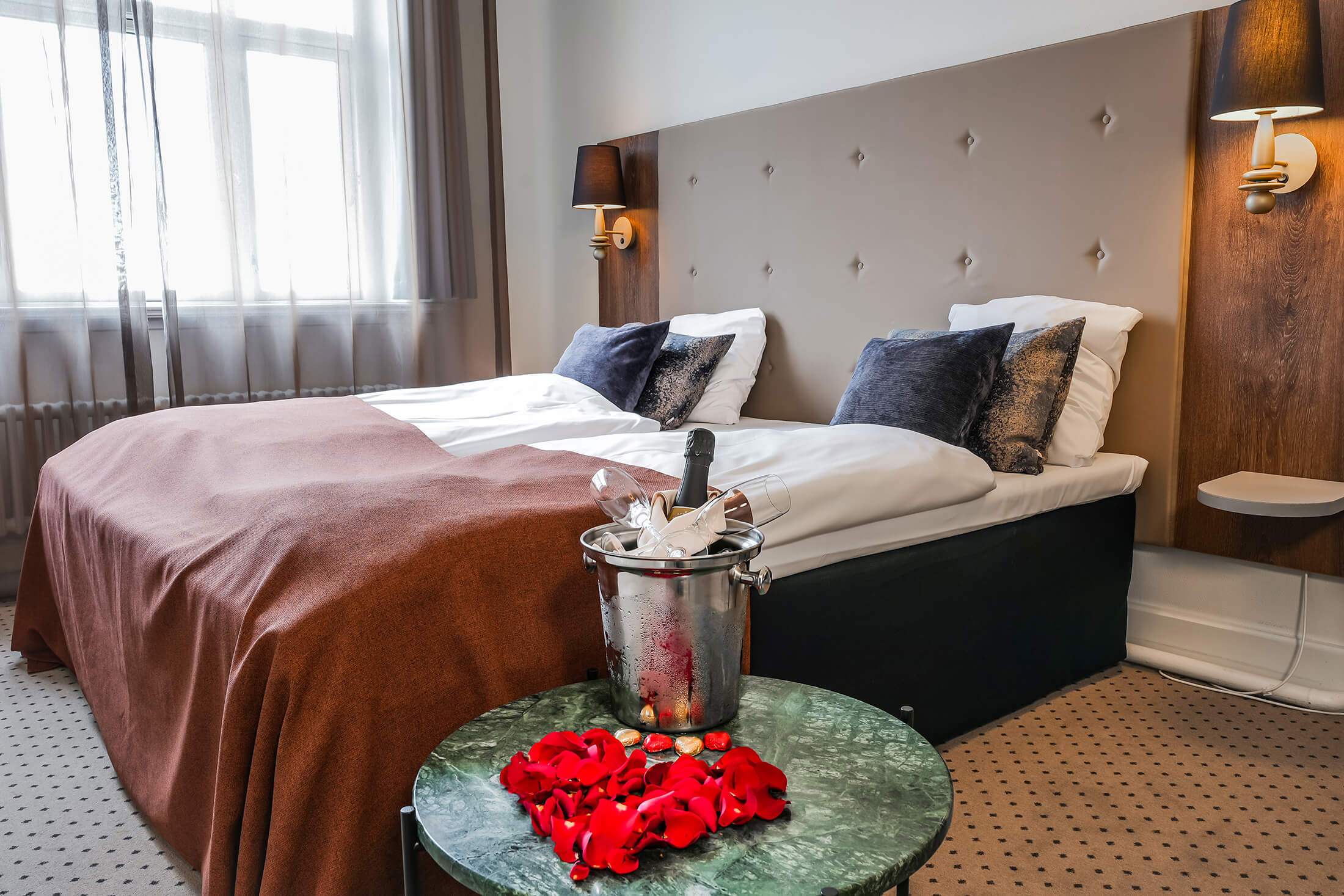 Et hotelværelse hvor der foran sengen står et bord med vinkøler og rosenblade