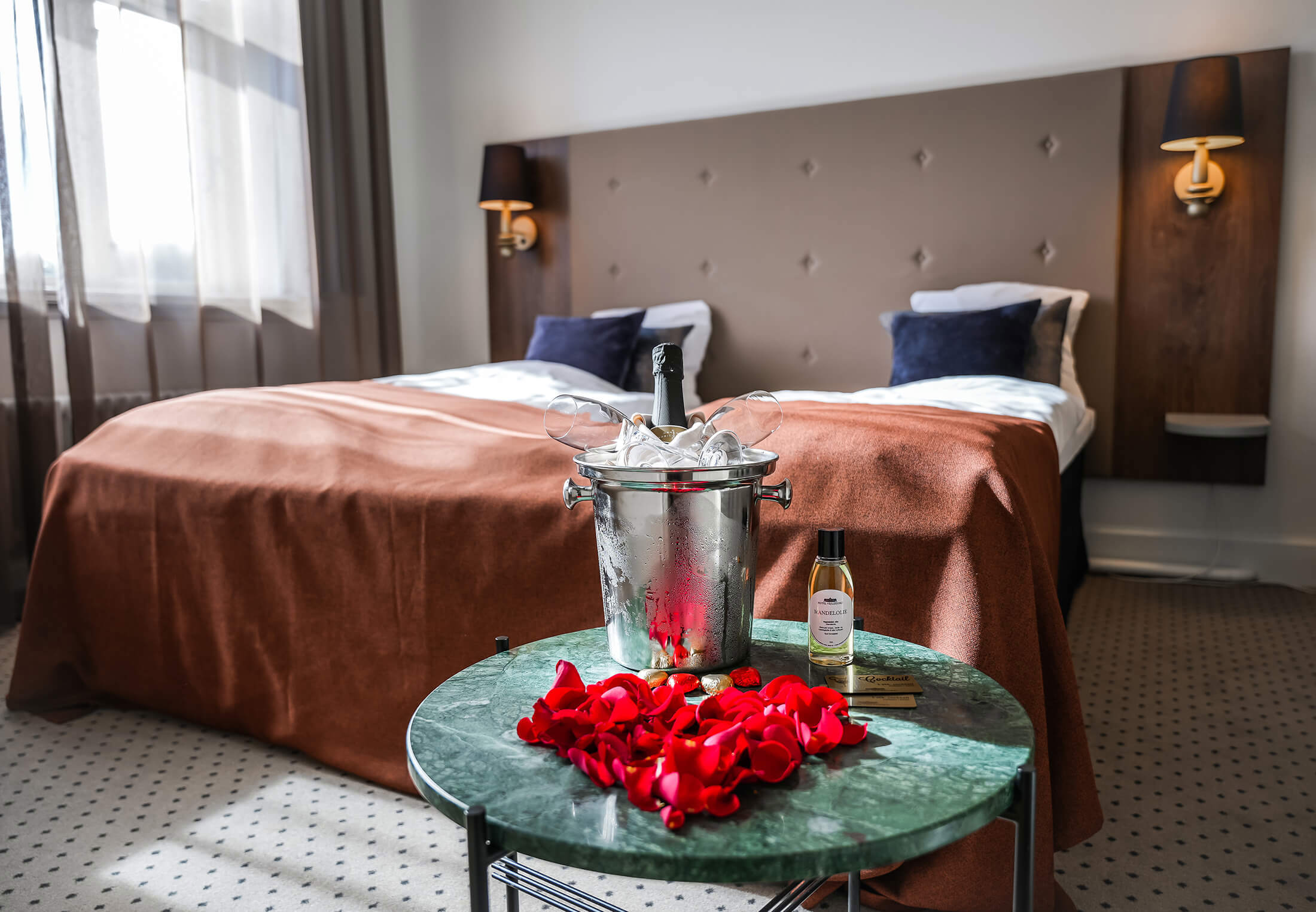 Et hotelværelse hvor der foran sengen står et bord med vinkøler og rosenblade